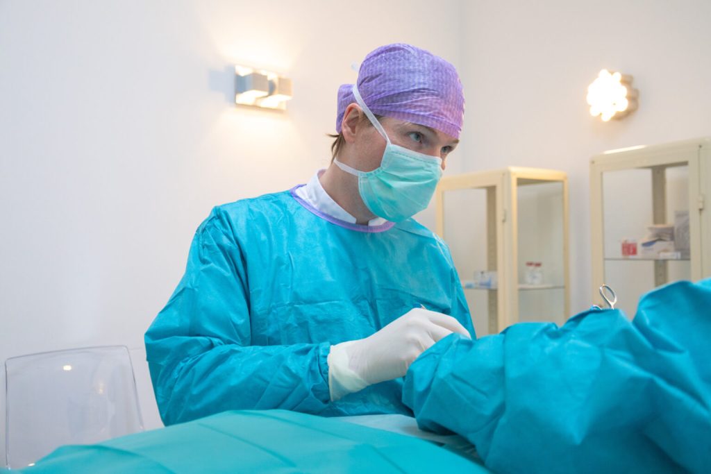 Clinician in a blue surgical gown, face mask and purple cap working in a treatment room.